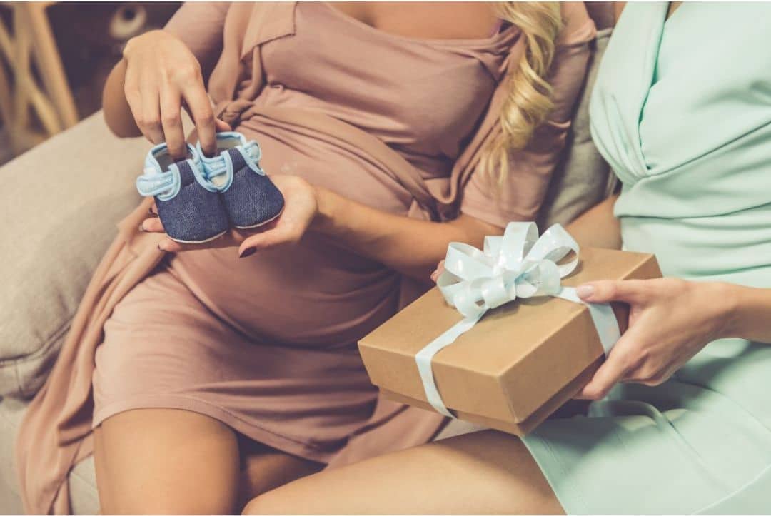 Schwangere Frau in beigem Kleid mit kleinen blauen Babyschuhen in der Hand, danaben eine weitere Frau mit einem Geschecnk mit blauer Schleife. Baby Party baby shower